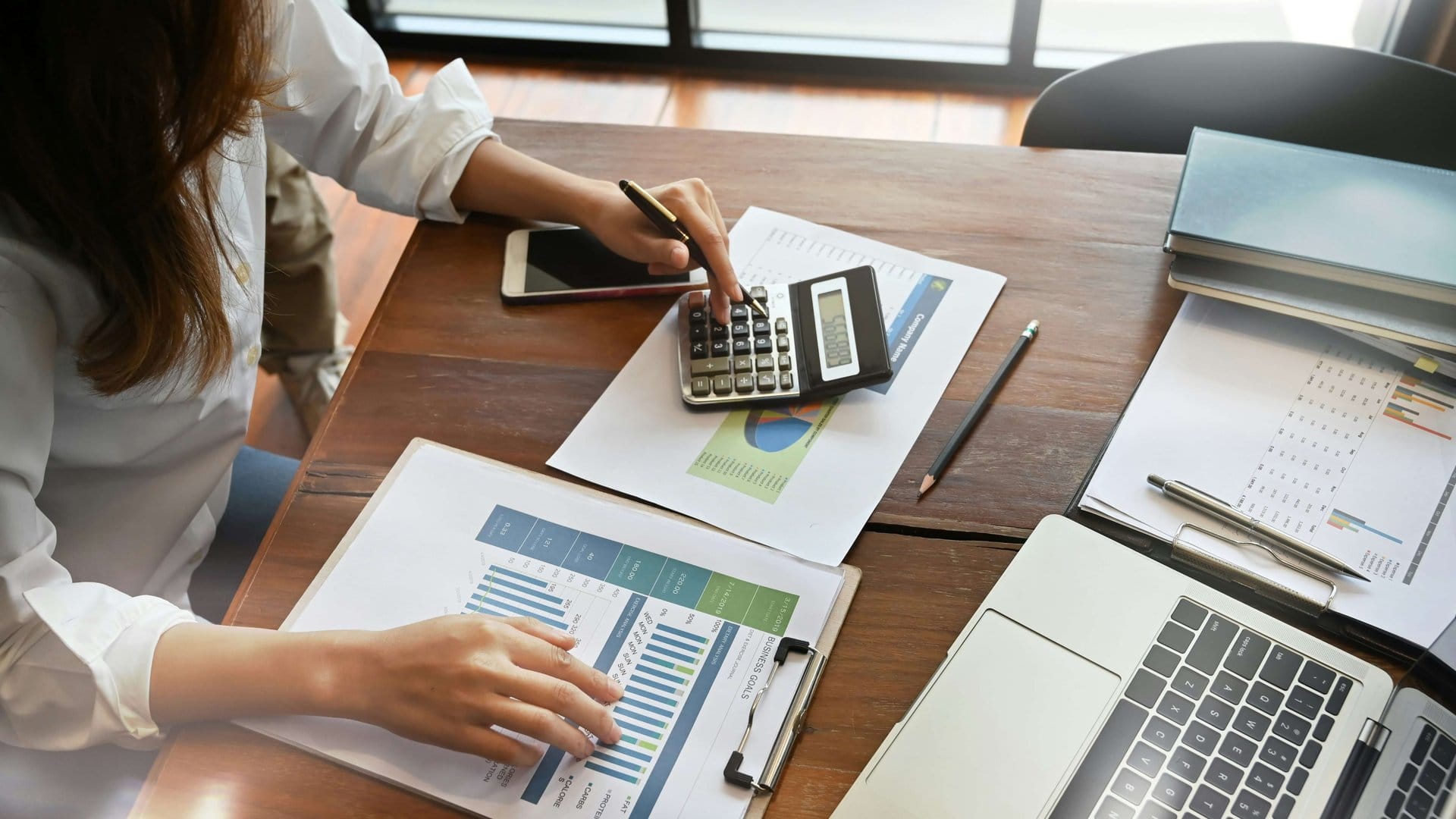 Person using a calculator on a wooden desk covered with charts and papers