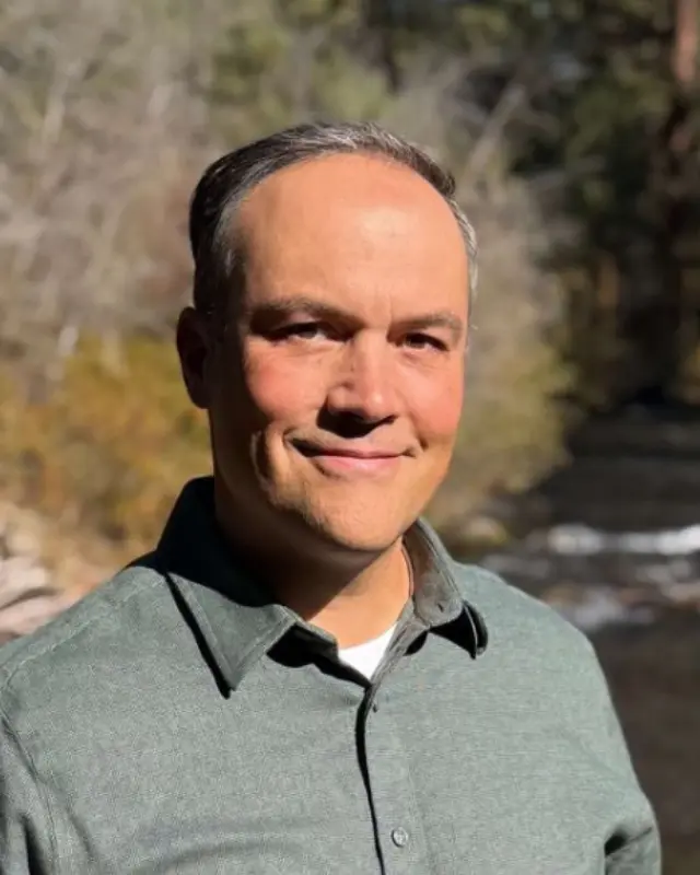 Close up of Mike Zacha smiling outdoors in a green button-up shirt with a river behind him